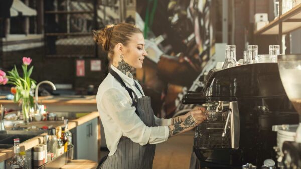 Eine Frau mit Schürze arbeitet in einem Cafe und bedient einen Kaffeeautomaten.
