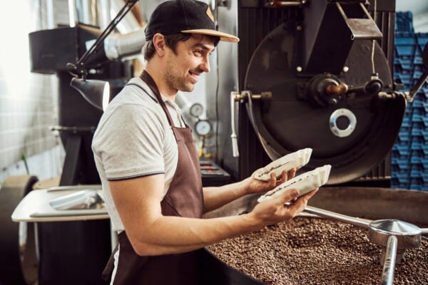 Un homme portant un tablier et une casquette vérifie en souriant la qualité des grains de café fraîchement torréfiés dans un torréfacteur.